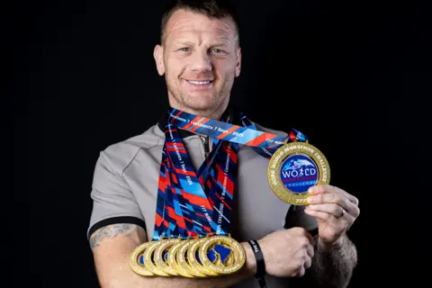 World Marathon Challenge A grinning Bobby, who is wearing a grey t-shirt, balances six of his medals on the right forearm and holds aloft another with his left hand