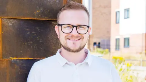 Brendan Hall A man with ginger hair, a small ginger beard, black glasses and a white polo shirt stands in front of some newbuild houses. They are blurred in the background. He is smiling.