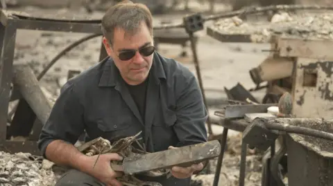 Peter Mitchell has a short beard and brown hair, with a dark shirt. He sits looking at the remnants of a piece of furniture - surrounded by the burnt out shell of his home