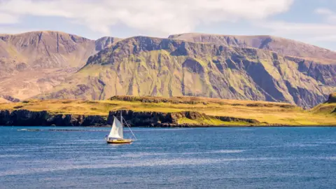 A yacht with a white sail cruises past Rum, and the island's rugged coastal hills.