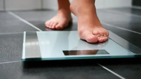 Person weighing themselves on scales. A set of scales can be seen on the floor of a bathroom. The feet of a person is in view as they step on the scales.