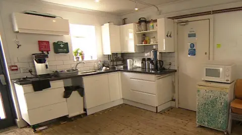 A kitchen with white cupboards on bare floorboards. It has exposed pipework and walls which need repainting.