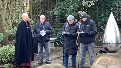 Luke Deal/BBC Reverend Canon Matthew Vernon stands and looks on at two other gentleman speaking into microphones during the Holocaust Memorial Day event. They were coats and the two men speaking wears hats as they read pieces of paper in their hands.