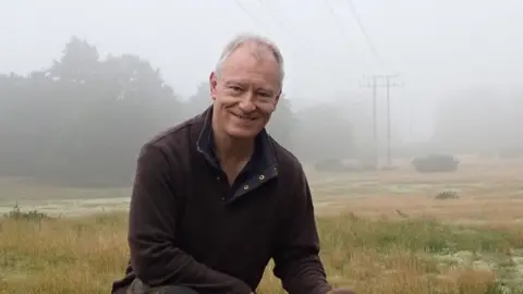 Bill Amos Bill Amos wearing a brown sweatshirt, smiling broadly and crouching on grassland on a misty day with telegraph poles in the distance