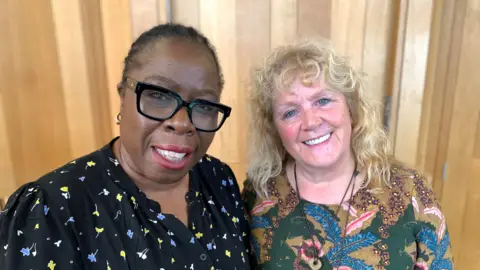 Two women stand next to each other, smiling at the camera. The woman on the left has black hair, scraped back, and is wearing a black shirt with yellow and blue flowers on it, and black glasses. The woman on the right has long curly blonde hair and is wears a colourful patterned top.