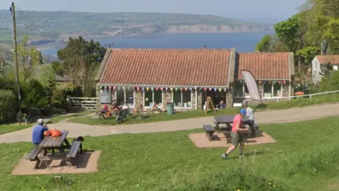 Google A small stone café with a red‑tiled roof sits on a grassy coastal hillside, decorated with colourful bunting, as people sit at picnic tables or walk and cycle nearby, with the sea and distant cliffs visible behind it.