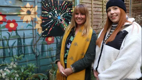 Potteries Museum & Art Gallery Prof Claire Gwinnett and Laura Wilkinson are stood outside by a green painted fence next to artwork. Claire has long brown/blonde hair and is wearing a large yellow scarf over a dark jacket and patterned top. Laura has a white jacket on over a top with long, red hair and is wearing a bobble hat. 
