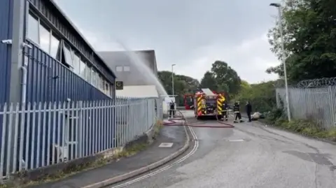 Fire engine with a firefighter using a red hose to extinguish the fire at Pixon Trading Centre in Tavistock