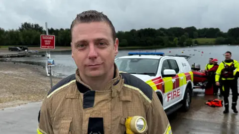 A firefighter stands by the side of a lake, with a fire service vehicle in the background