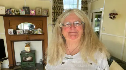 Shaun Whitmore/BBC Di Cornell who has below shoulder-length fair hair, rectangular clear-freamed glasses and is wearing a white top. She is standing in her sitting room. Behind her is a wooden fireplace, its mantle covered in cards, and a green wood burner-style heater. 