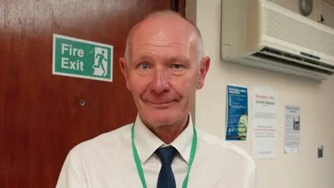 Darryl Preston head-and-shoulders photo, he is wearing a white shirt, blue tie and green lanyard.