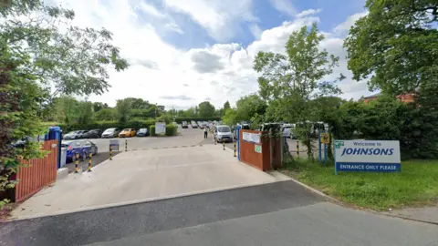 A Google Streetview image of the entrance to a car park. Lots of cars are parked inside. A sign at the entrance, which leads off a road and has grass and trees to the side, reads: "Welcome to Johnsons."