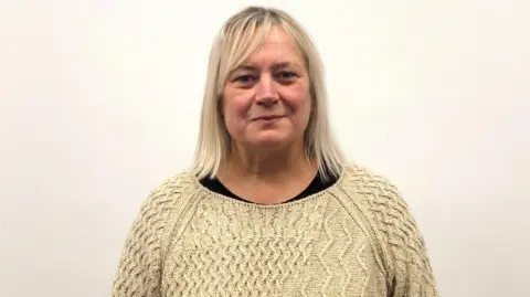 Caroline Doherty standing against a plain white wall, wearing a light-yellow knitted sweater over a dark top. She is centred in the frame from the shoulders up. She has blonde shoulder length hair.