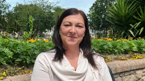 A woman with dark brown hair sits on a bench in front of a brick wall. Greenery, plants, and trees are behind her, with some smaller yellow flowers. She wears a white, thin jumper.