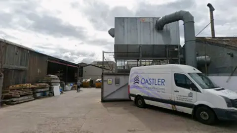 The entrance and yard of a construction company. A van is in the foreground with the name Oastler on the side. Pallets and wood are in the yard