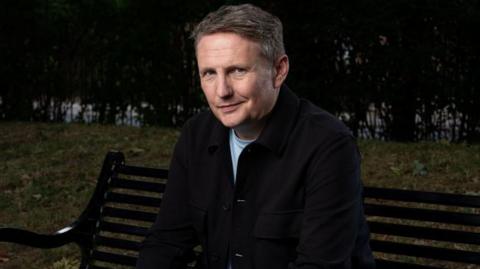 Mark O'Sullivan sitting on a bench. He is wearing a black jacket over a white t-shirt.