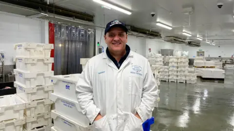 A man wearing a white shirt stands inside warehouse. He has his hands in his pocket and is wearing a blue cap. White boxes are stacked on top of each other around him. Strip lights are turned on. 
