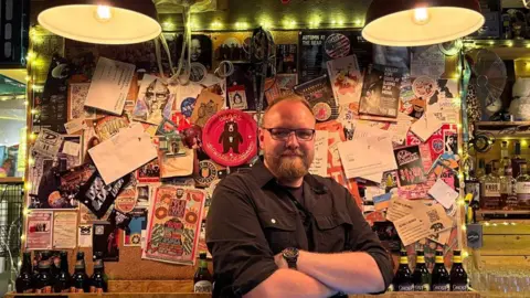 Karl Brown wearing glasses and a dark button-up shirt stands with arms folded behind a bar or music venue counter. Behind the person is a large noticeboard densely covered with gig posters, flyers, stickers, postcards and notes, illuminated by warm string lights. Two large industrial-style hanging lamps dominate the top of the scene. Bottles of beer and spirits are arranged on shelves and counters around the space, creating a cosy, eclectic atmosphere filled with music and pub memorabilia.