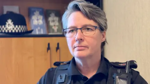 Louise Kennaugh has short dark and grey hair and wears glasses. She's in black police uniform sitting in an office, with a wooden cabinet behind to the left with her police hat resting on a shelf. 