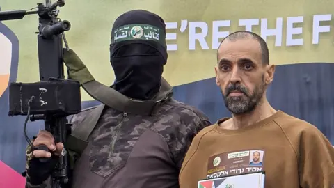 A masked Hamas fighter with Israeli hostage Eli Sharabi  on a stage before he was handed over to a Red Cross team in Deir el-Balah