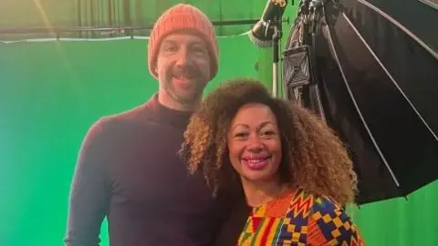 Andy Johnson wears an orange hat and a black rollneck jumper. He has his arm around his wife Ithalia who has long curly hair and wears a multi-coloured top. They are standing in front of a TV green screen and are smiling at the camera.