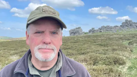 Alan has a white beard and is wearing a cap. He is standing in front of Hound Tor. A pile of granite rocks can be seen just over his shoulder in the background.