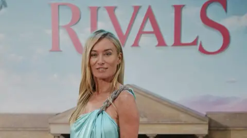  Victoria Smurfit arriving for the world premiere of season two of Rivals, at BFI IMAX in Waterloo, central London. She has long blonde hair, wearing a light blue gown. A sign for Rivals is behind her.