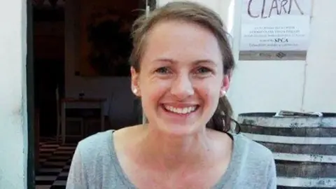 Head-and-shoulders portrait of a person with hair tied back, wearing pearl stud earrings and a light-coloured top, facing the camera indoors.
