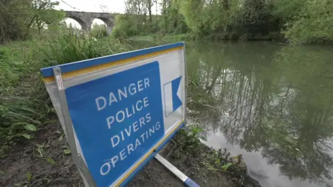 A sign which says "danger police divers operating" which is next to a river. It is a blue sign with white writing on it. There are bridge arches in the background which are from the railway bridge. 
