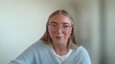 A women with dark blonde hair wearing pink glasses, v-neck sky blue jumper and a white shirt. The background is blurred. 