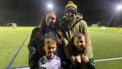 Sophie and Hannah wrapped up warm, standing with their children wearing sports kits. 