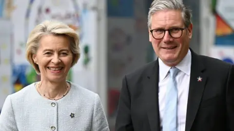 A man and woman look towards the camera. The man is wearing a dark suit, white shirt and light blue tie. He has grey hair and is wearing dark rimmed glasses. The woman has short blonde hair and is wearing a light grey buttoned jumper. Both are wearing badges in the shape of a small star.