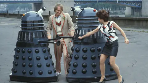 A young man in white cricketing gear and a young woman in a black and white patterned top and a black leather mini skirt attacking two grey Daleks on the banks of the River Thames - taken from Doctor Who filming in 1983