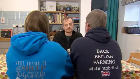 Punggung seorang pria dan seorang wanita duduk di meja dapur berbicara dengan seorang reporter BBC. Mereka mengenakan hoodies yang mengatakan 'Back British Farming' dan 'hanya mencoba menyatukan domba saya' di punggung. 