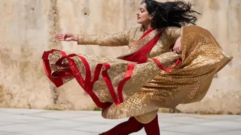 Sonia Sabri Sonia is mid-performance with traditional dress on. Her clothing is gold and red and floats in the air as she swirls and turns.
