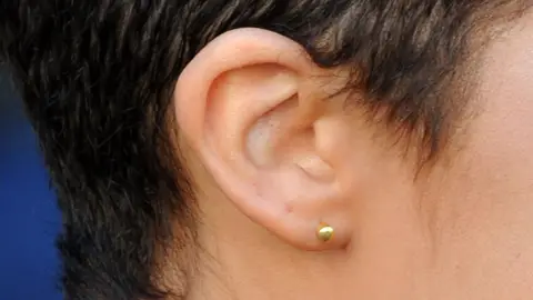A side view of a woman's ear, the lobe of which is pierced with a gold stud. She has short dark hair.