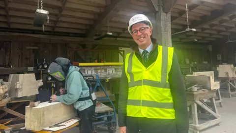 BBC/ Emily Johnson A man in a hi-vis jacket and helmet standing next to a female stonemason  chiselling a block of stone 