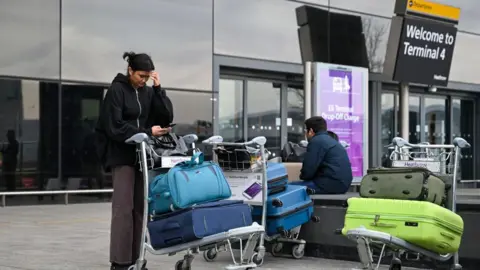 Getty Images Woman and man wait with brightly coloured suitcases on trolleys outside Departures at Heathrow Terminal 4