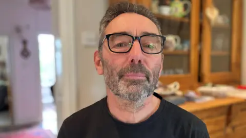 Wearing glasses and a black T-shirt, Peter Holden poses for a photograph in his kitchen at his Worcestershire home. He has the start of a beard with dark and white hair.
