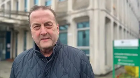 Phil Bialyk in a blue quilted jacket standing outside the Civic Centre offices of Exeter City Council against a blurred background.
