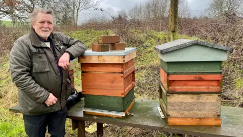 BBC A man stands beside three homemade beehives in the middle of a field. The man has long grey hair, a thick green coat and black trousers. He is resting his elbow on one of the hives which has three bricks on top of it. The hives sit on a wide wooden bench. Behind the hives is a wire fence held up with wooden poles. In the distance is a large field with trees and shrubbery.