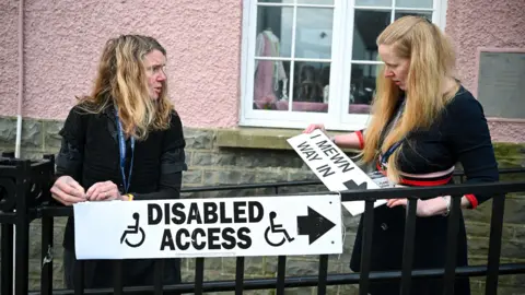 Finnbar Webster / Getty Images Two people erect signs outside a pink building in St Davids, Wales on Thursday morning, one saying 'disabled access' with an arrow pointing right, and the other saying 'I mewn, way in', also with an arrow.