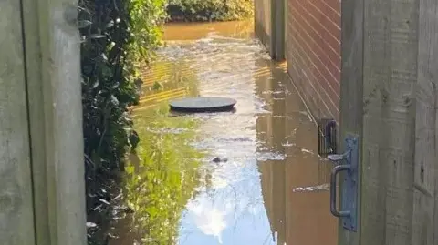 An image of a risen manhole cover and raw sewage water flooding out from underneath along a path around flats
