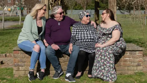Hospice in the Weald Three women and one man sitting on a brick bench at a park. There are trees behind them. 