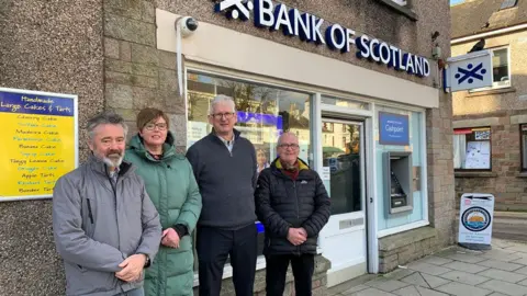 A concerned group of residents - three men and one woman - stands in front of the Moffat Bank of Scotland branch