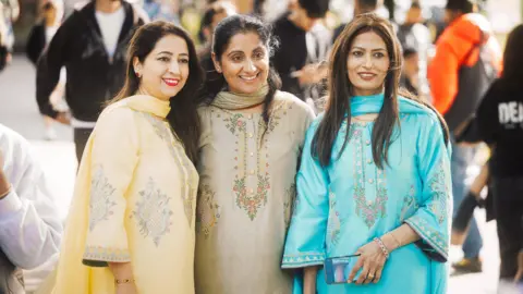 Three Asian women, all dressed in brightly-coloured dresses of yellow, grey/green and turquoise, smile for the camera while outdoors at the festival