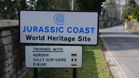 BBC A road sign with the words "Jurassic Coast World Heritage Site" on it with a sign below it with the words "Twinned with Norden, Sully-Sur-Loire and Elblag" with flags alongside