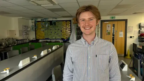 Hazel Shearing / BBC Daniel is seen standing in a science classroom or laboratory, wearing a striped shirt. Long workbenches with taps and stools fill the room, while a noticeboard covered with coloured papers and a door marked with a fire exit sign are visible in the background.