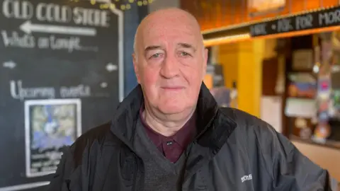 A man wearing a grey knitted jumper and burgundy shirt under a black coat. He is standing in front of a bar. There is also a chalkboard behind him.