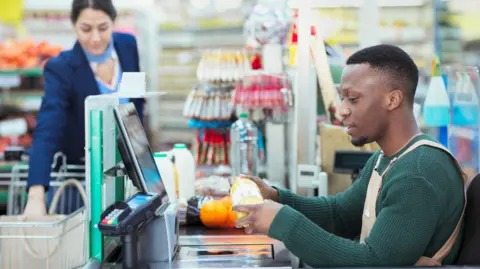 Getty Images Young male shop assistant in a green top and cream overall sits at a till and scans a bottle, while a female customers loads food and drink products onto the conveyor belt. 